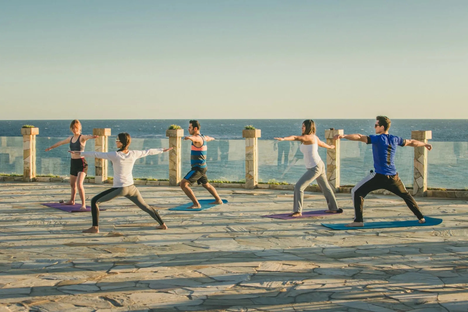 Group holding a warrior yoga pose on a stone terrace above the ocean