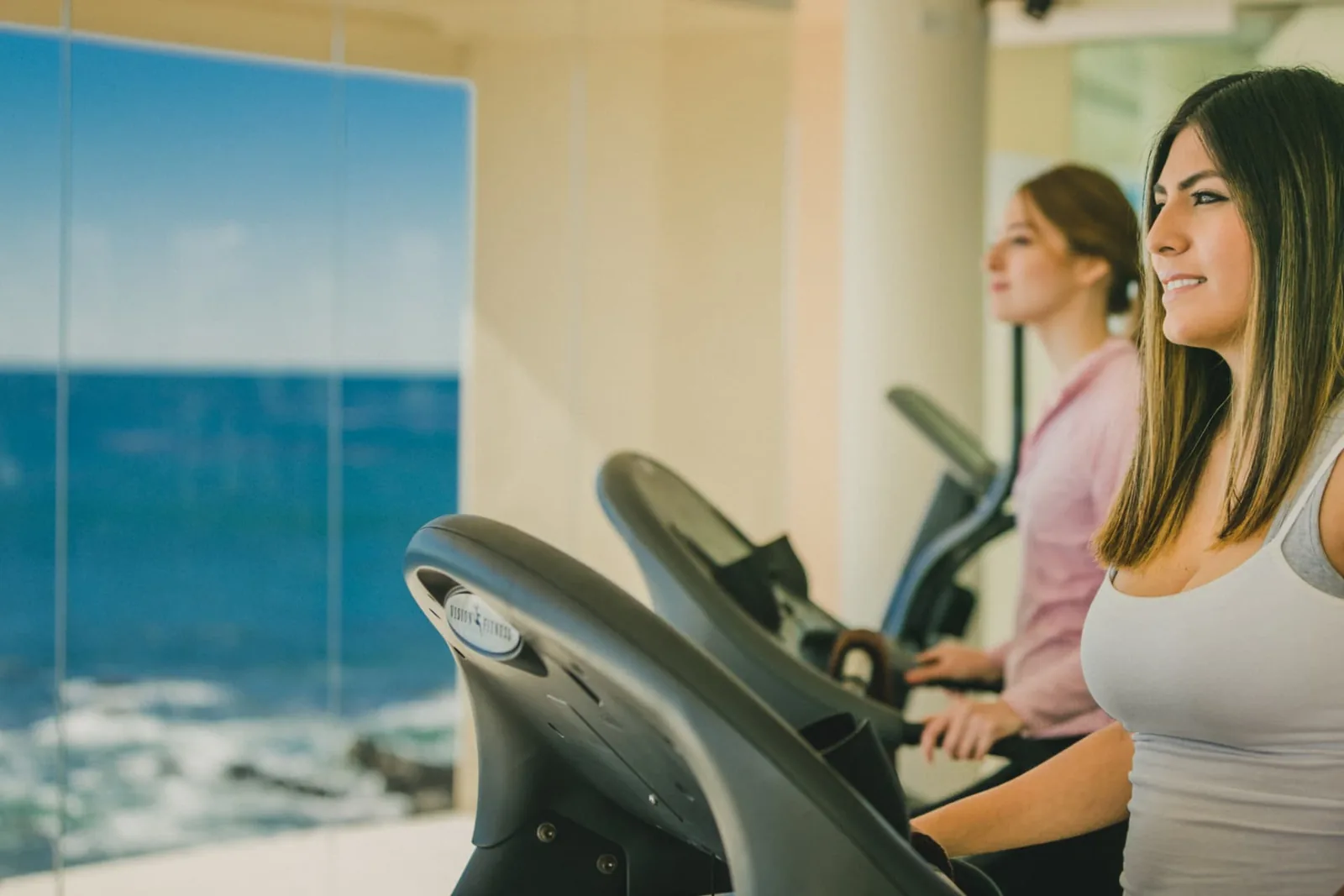 Two women using cardio machines beside a window overlooking the ocean