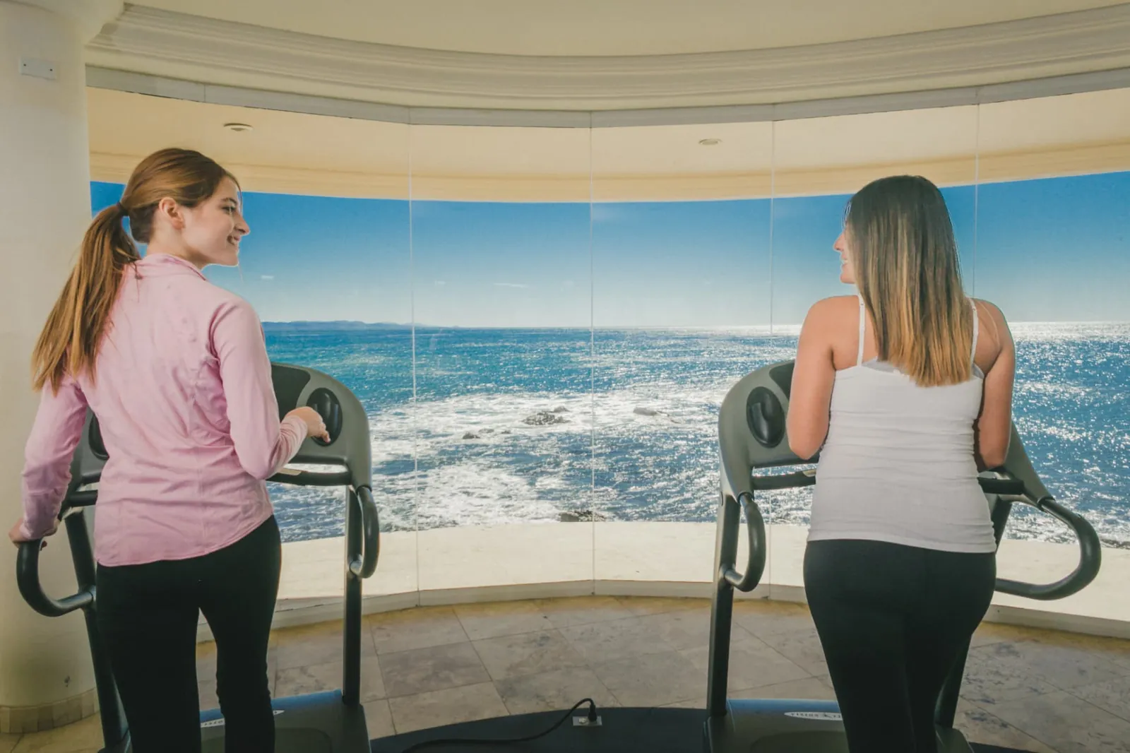 Two residents walking on treadmills in front of a curved ocean-facing window